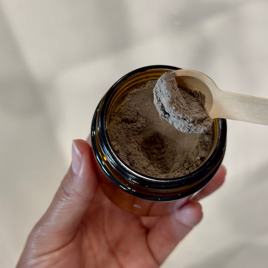 A hand holding an open amber glass jar filled with a dark brown powder, with a small wooden spoon lifting some of the powder above the rim.
