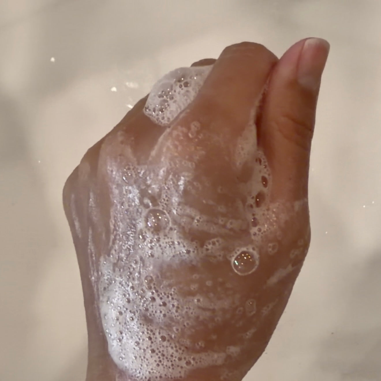 A close-up of a hand covered in lathered soap foam, with fingers curled together, against a soft beige background.