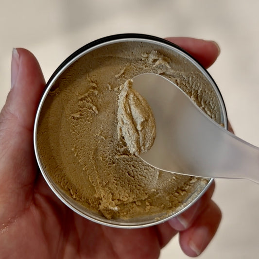 A hand holding an open round metal tin filled with a beige cream, with a translucent spatula resting in the product against a soft neutral background.