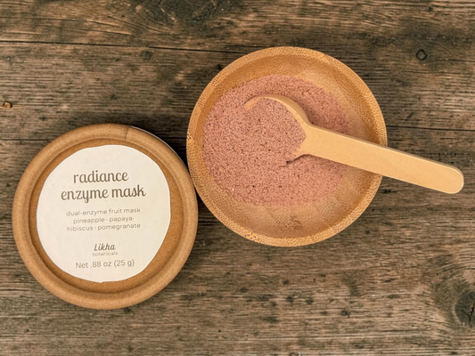 Radiance enzyme face mask in round tin beside wooden bowl filled with pink-coral powder and bamboo scoop on wood surface