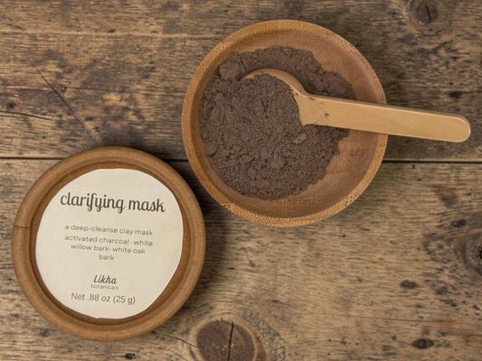 Clarifying powdered face mask in round tin beside wooden bowl filled with dark charcoal powder and bamboo scoop on wood surface