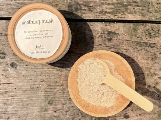 Soothing powdered face mask in round tin beside wooden bowl filled with cream-colored powder and bamboo scoop on wood surface