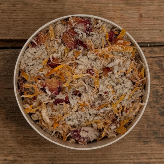 Botanical bath tea in round container filled with dried botanicals including lavender, calendula petals, rose petals, and oats on dark wood surface