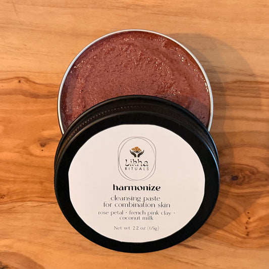 An open round metal tin of Harmonize Cleansing Paste, showing the textured pinkish-brown product inside, with the black-lidded container and white label in front, set on a wooden surface.