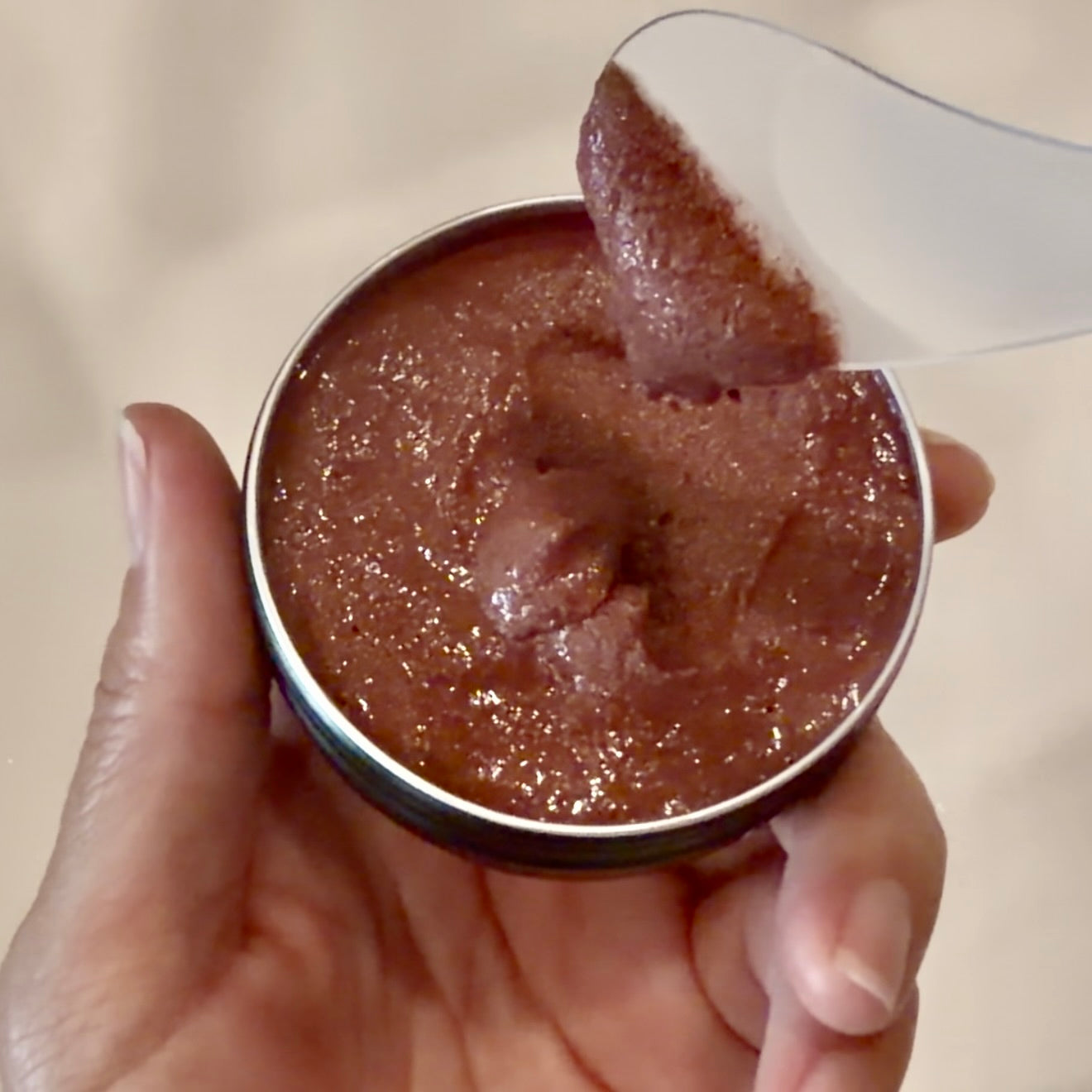 A hand holding a small round metal tin filled with a reddish-brown textured paste, with a white spoon lifting some of the substance.