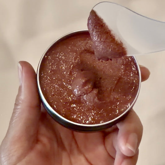 A hand holding a small round metal tin filled with a reddish-brown textured paste, with a white spoon lifting some of the substance.