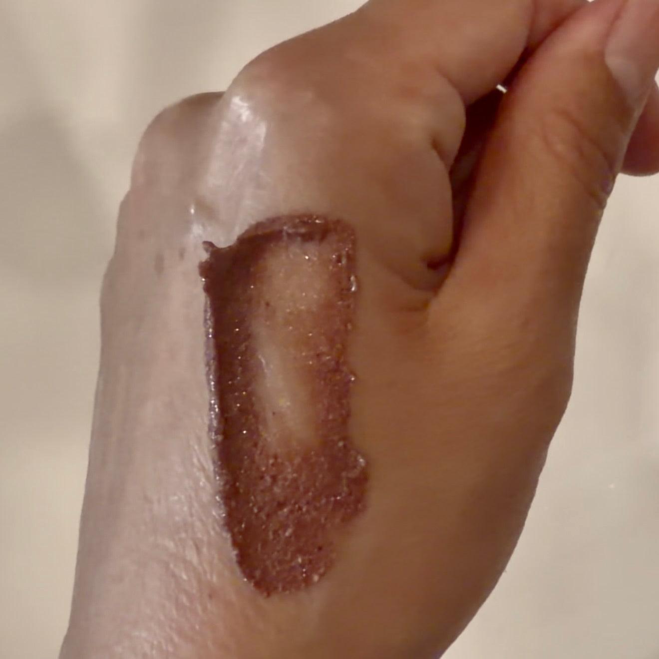 A close-up of a hand with a swatch of shimmering brown cosmetic product applied in an irregular vertical streak on the back of the hand.
