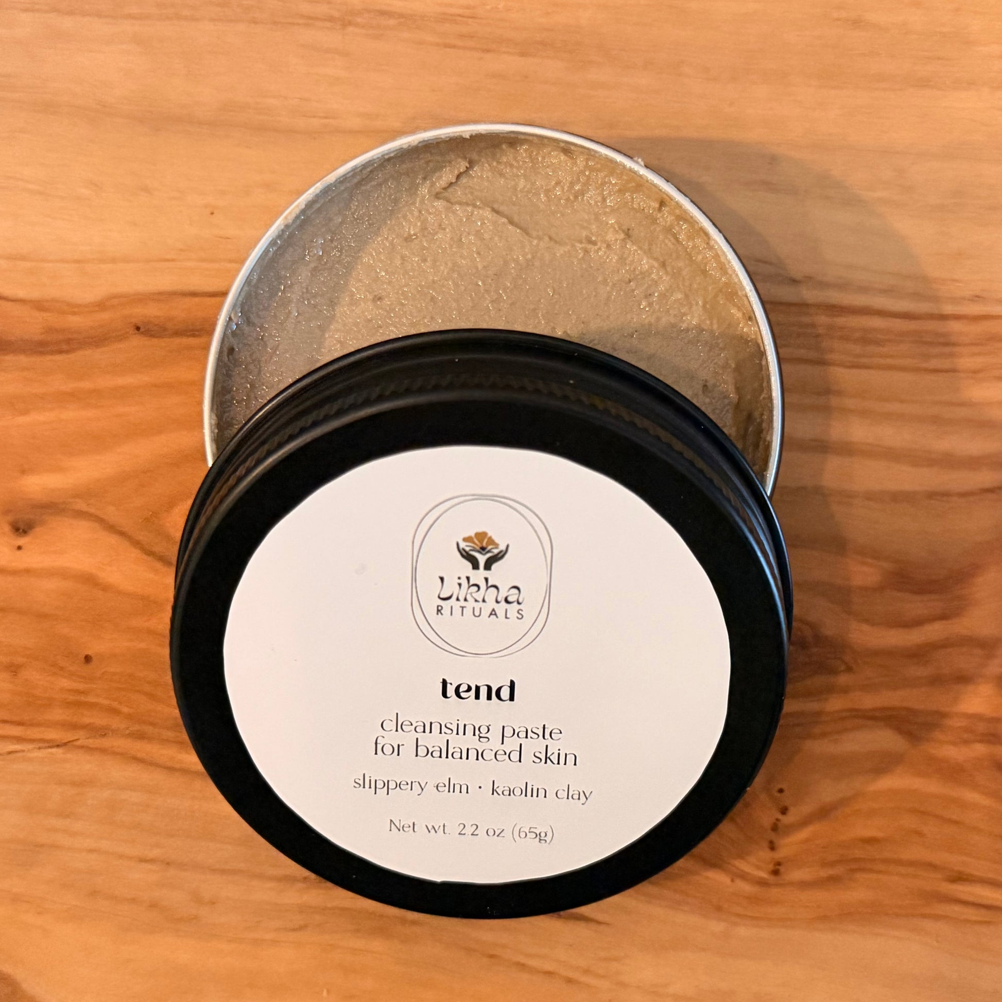 An open round metal tin of Lisha Rituals 'Tend' cleansing paste sits on a wooden surface, with the black-lidded jar placed in front showing the white product label.